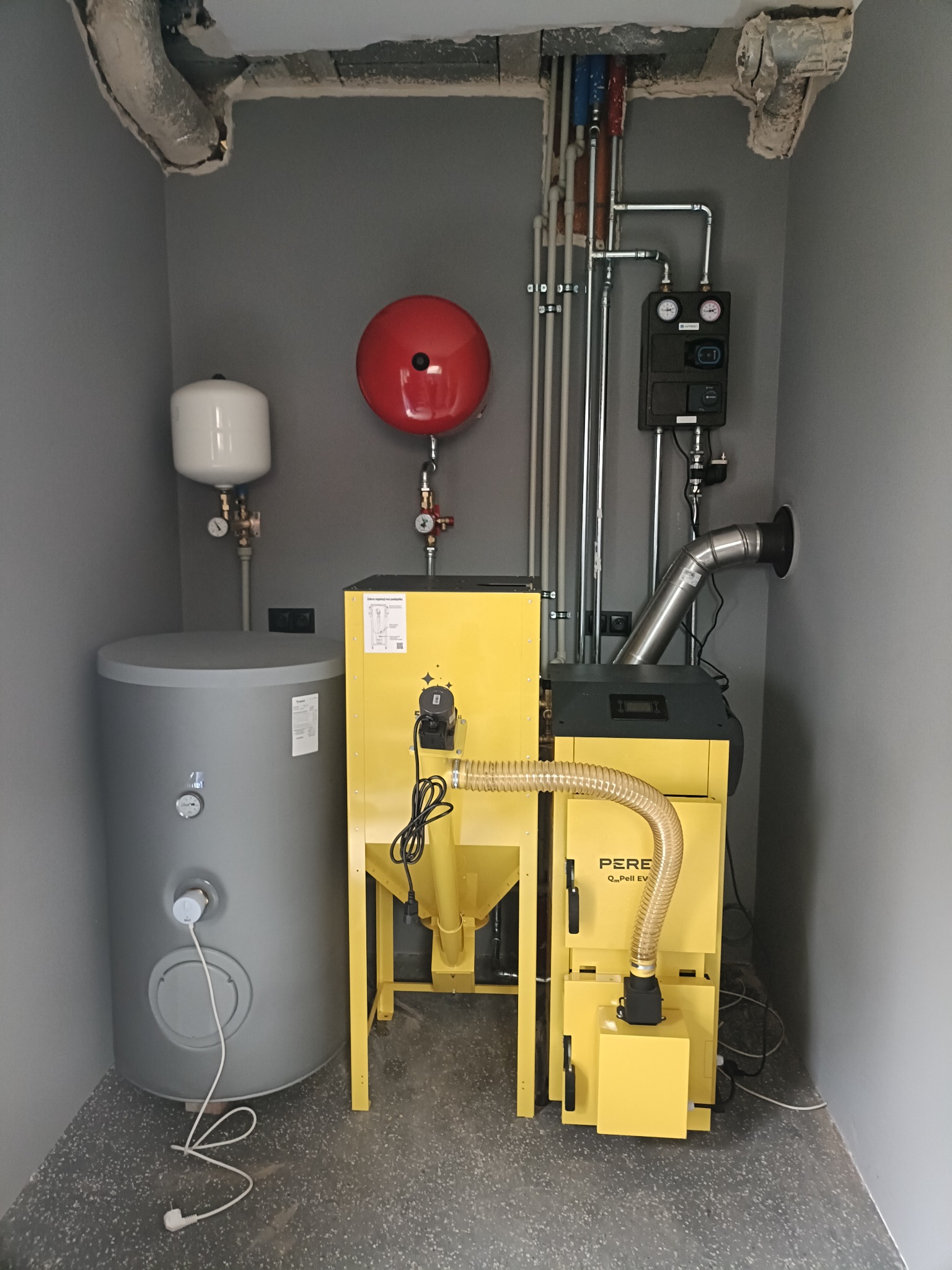 Żółty piec na pellet marki PEREKO, szary zbiornik na wodę i instalacja grzewcza w szarym pomieszczeniu. Widoczne rury, manometry i inne elementy instalacji.