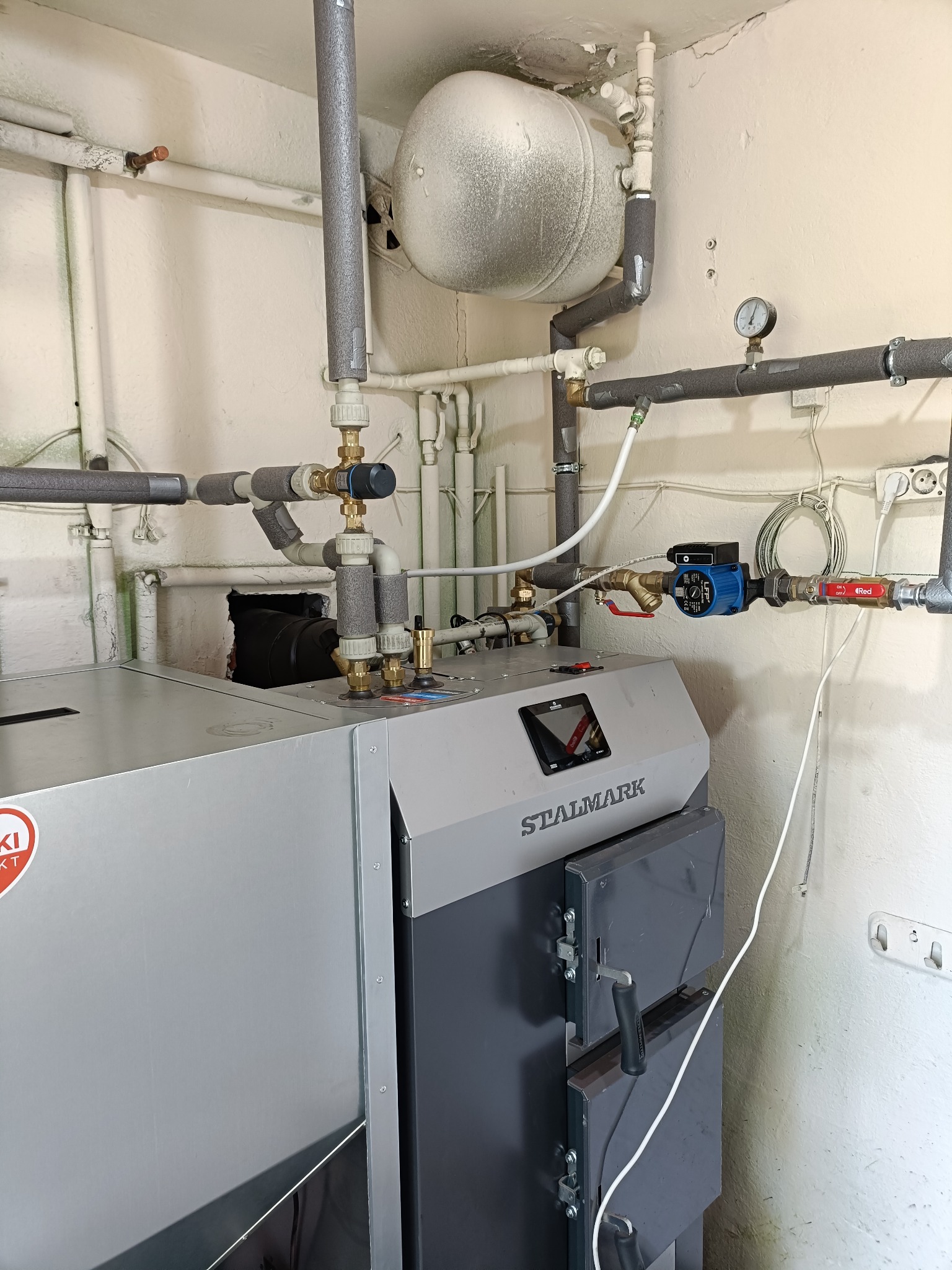 Szary piec CO marki Stalmark w kotłowni, z widoczną instalacją grzewczą: rury z izolacją, zawory, manometr i pompka obiegowa. Białe ściany, rury przy suficie.