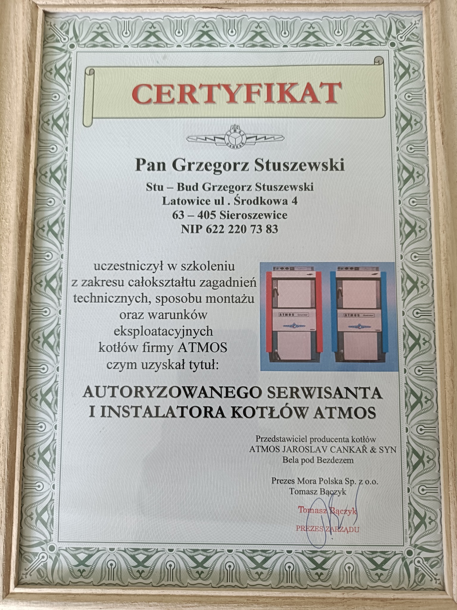 Certyfikat w ramce potwierdzający autoryzację serwisanta i instalatora kotłów Atmos, z danymi Grzegorza Stuszewskiego i adresem firmy. Widoczne zdjęcia kotłów.