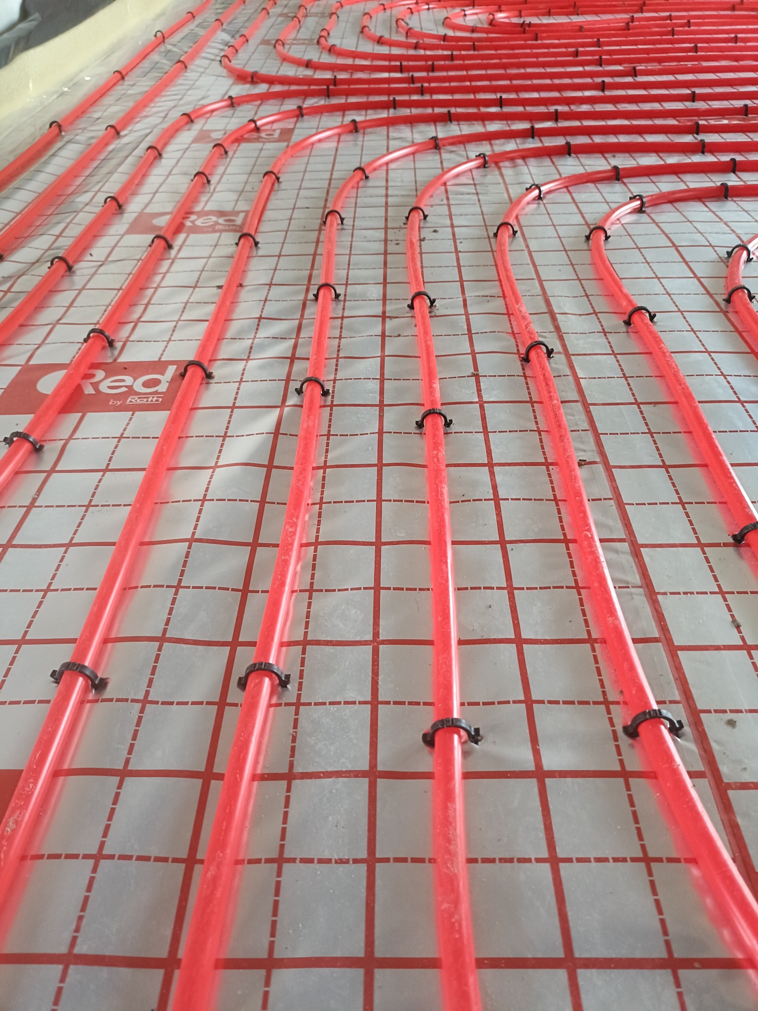 Czerwone rury ogrzewania podłogowego, mocowane czarnymi klipsami do folii z siatką. Widok z góry, ukośny kadr. Rury marki Red by Roth.