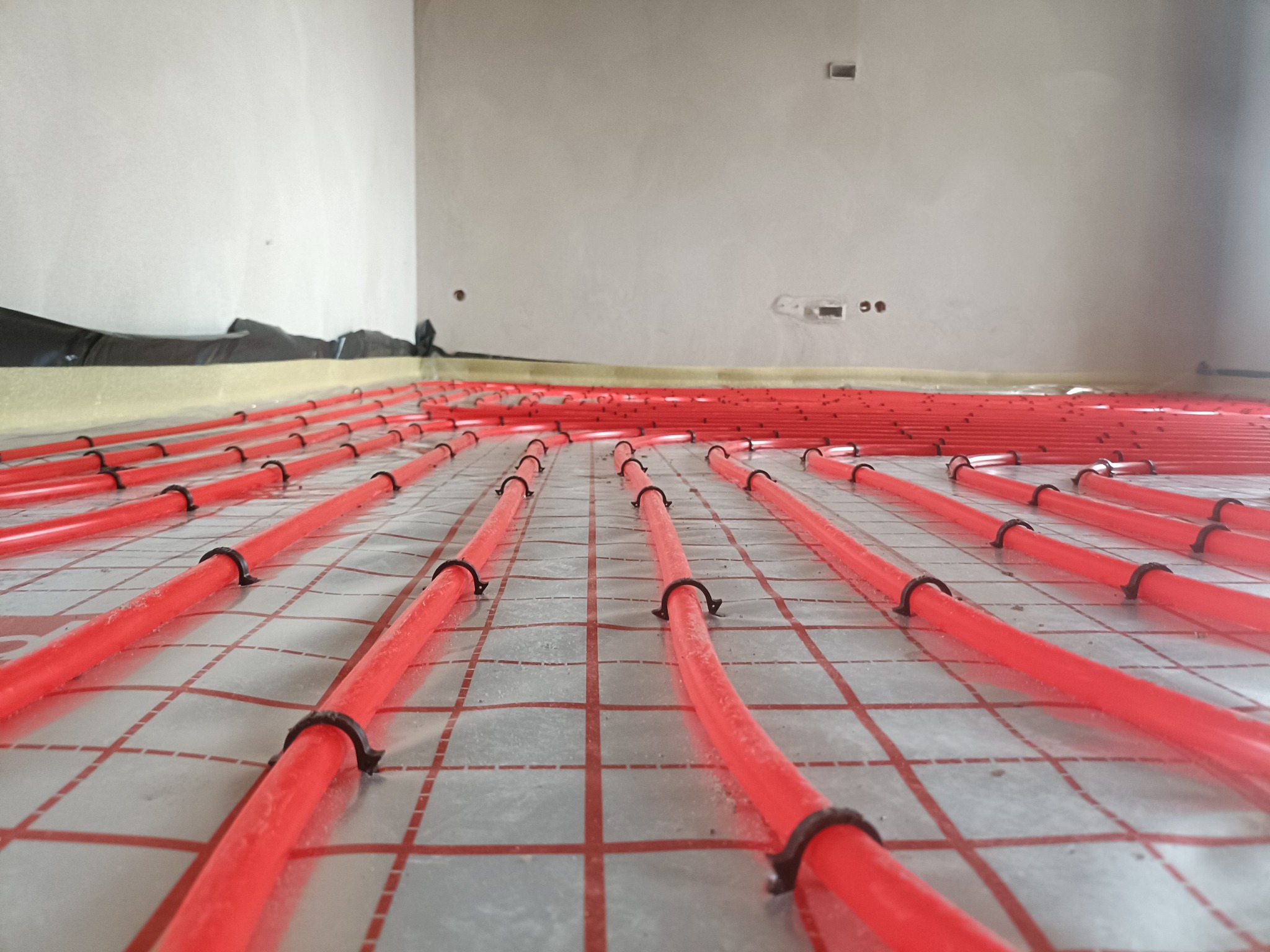 Czerwone rury ogrzewania podłogowego, równolegle ułożone na folii z siatką, przymocowane czarnymi uchwytami. Widok z niskiej perspektywy w surowym wnętrzu.