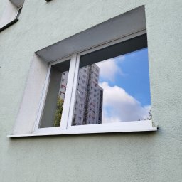 Waldemar Dondajewski - Okna w bloku z odbiciem budynku i błękitnego nieba z chmurami.