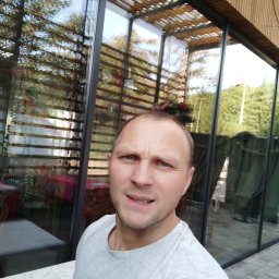 Waldemar Dondajewski - Mężczyzna pozuje na tle nowoczesnego budynku z dużymi, czystymi oknami, w których odbija się otoczenie.