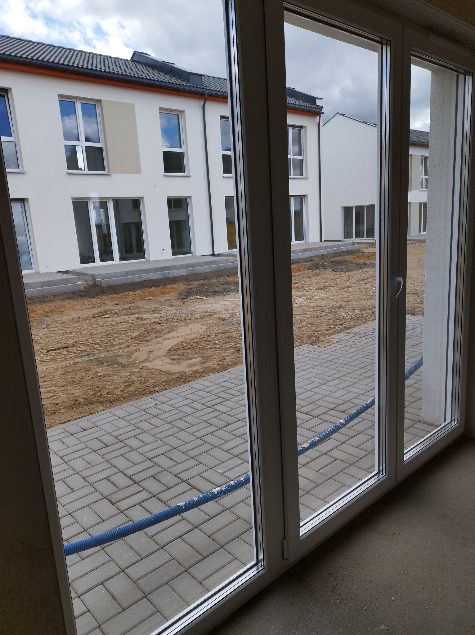 Widok z wnętrza przez nowe, białe okna na ułożoną kostkę brukową i budynki w tle, z niebieskim wężem na kostce.