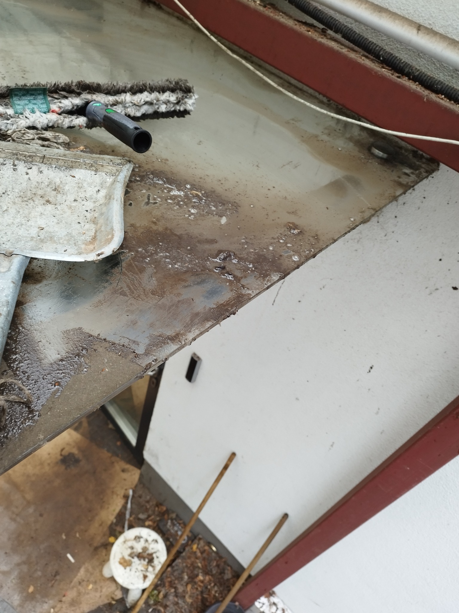 Mokry mop z szarą końcówką i skrobak na brudnej, metalowej powierzchni dachu nad oknem, widoczne ślady po czyszczeniu.