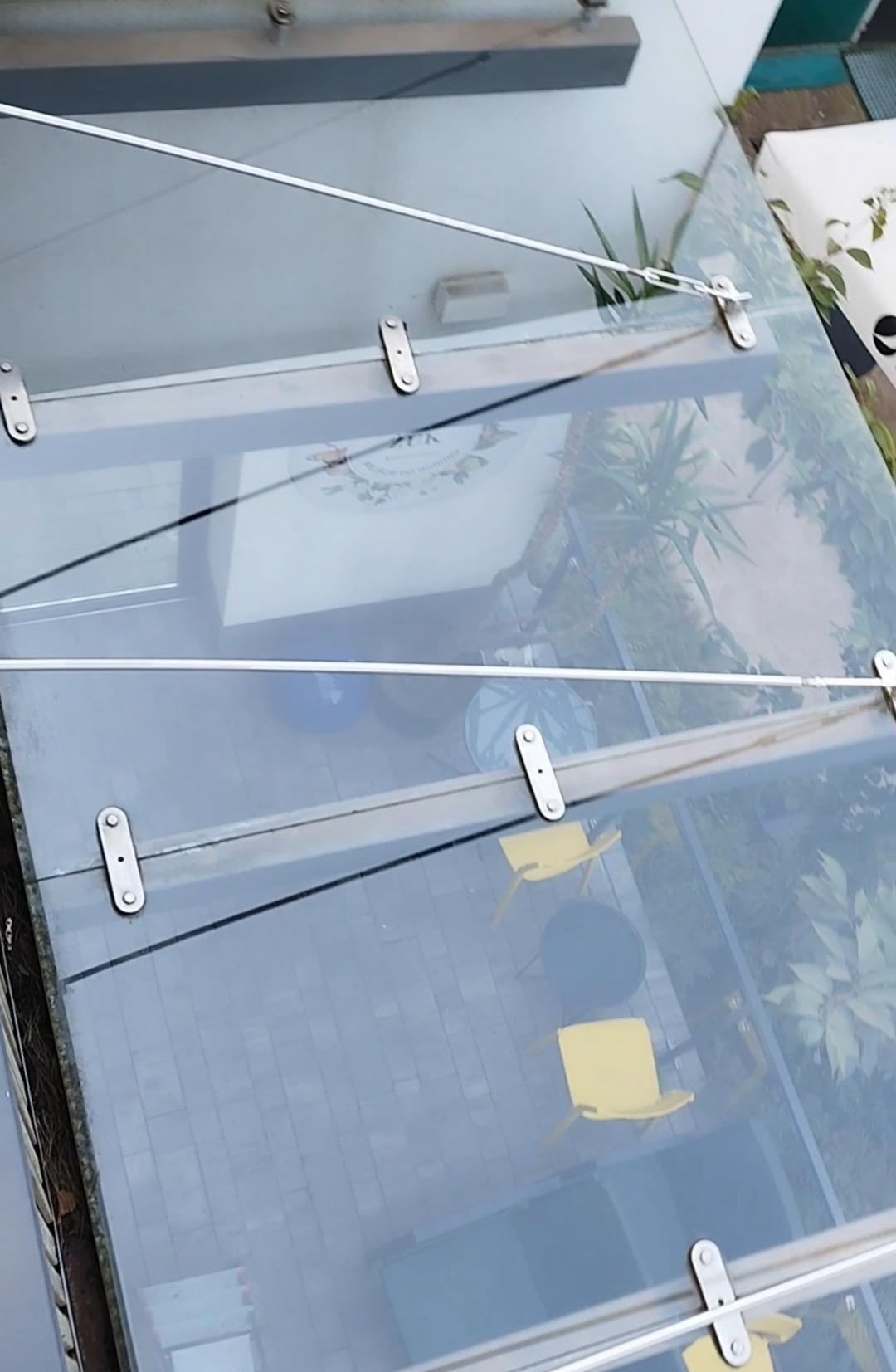 Przezroczysty szklany dach z metalowymi mocowaniami, widok z góry na patio z żółtymi krzesłami i stolikiem oraz roślinami.