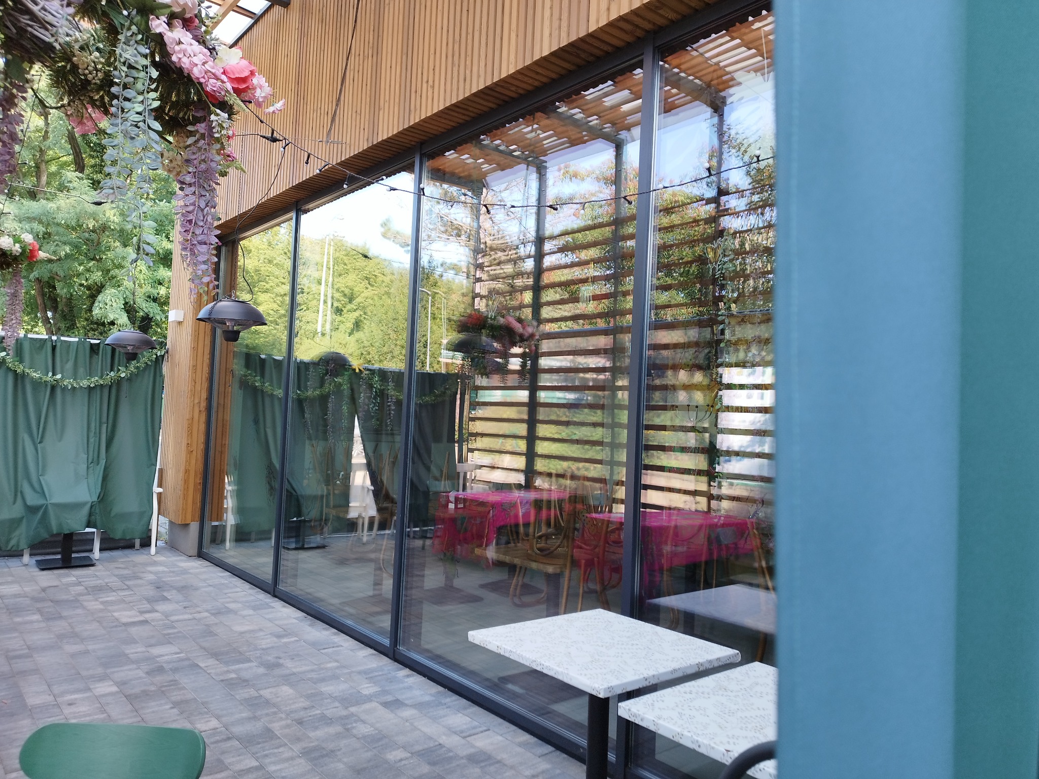 Szklana fasada restauracji z widocznymi stolikami wewnątrz i dekoracjami kwiatowymi na zewnątrz, odbijająca zieleń drzew.