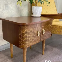 Stolik nocny po renowacji z geometrycznym wzorem, złotymi uchwytami i rośliną doniczkową. Widoczne odświeżone nóżki i żółty fotel w tle.