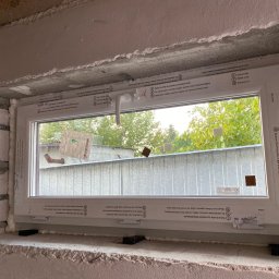 SERWIS I RENOWACJA „ANTA” - Nowe okno PCV z pianką montażową, folią ochronną i klinami poziomującymi, osadzone w surowym otworze okiennym z widoczną konstrukcją budynku w tle.