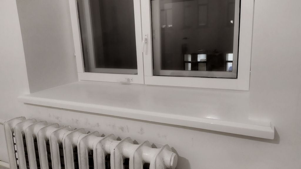 Białe okno PCV z widocznym uchwytem i parapetem, poniżej żeliwny, pomalowany na biało kaloryfer. Widok z bliska, wnętrze pomieszczenia.