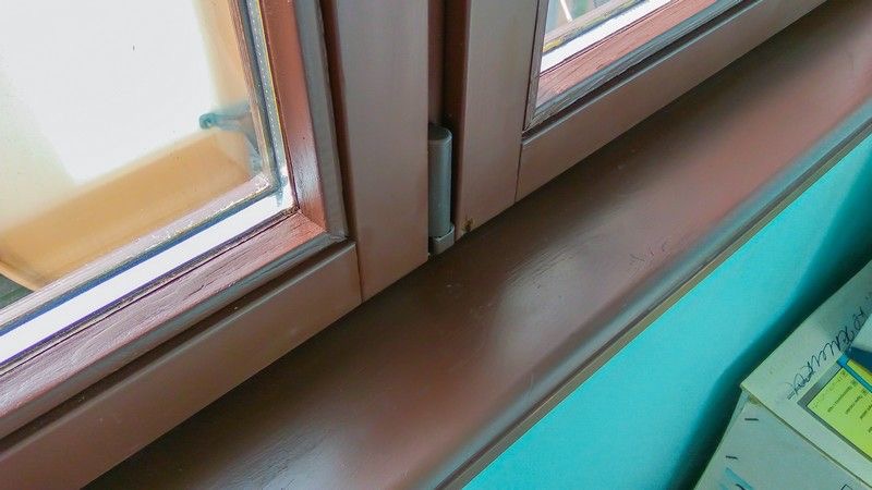 Brązowe okno PCV z widocznym zawiasem i parapetem na tle turkusowej ściany, ujęcie z bliska, detale konstrukcyjne.