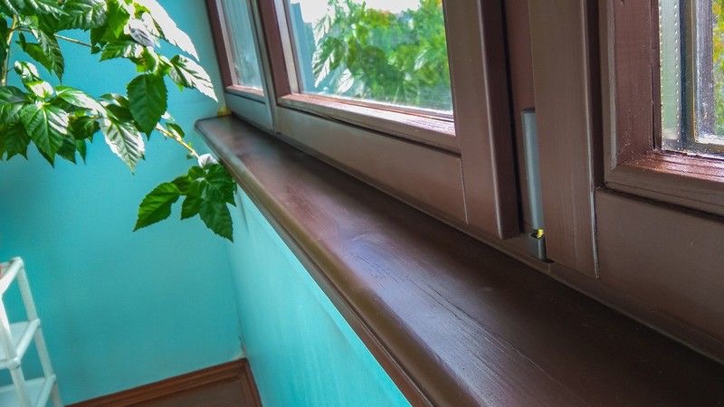 Brązowe okno PCV z widokiem na zieleń za oknem, z drewnopodobnym parapetem i fragmentem turkusowej ściany. Widoczny mechanizm okienny.