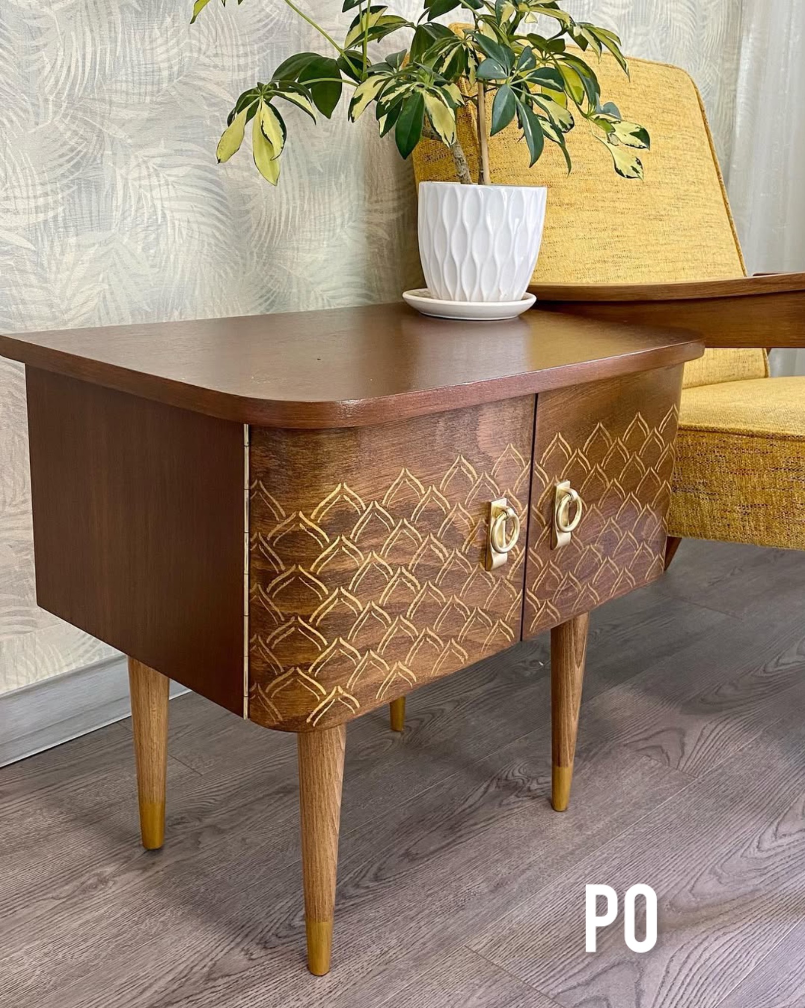Stolik nocny po renowacji z geometrycznym wzorem, złotymi uchwytami i rośliną doniczkową. Widoczne odświeżone nóżki i żółty fotel w tle.