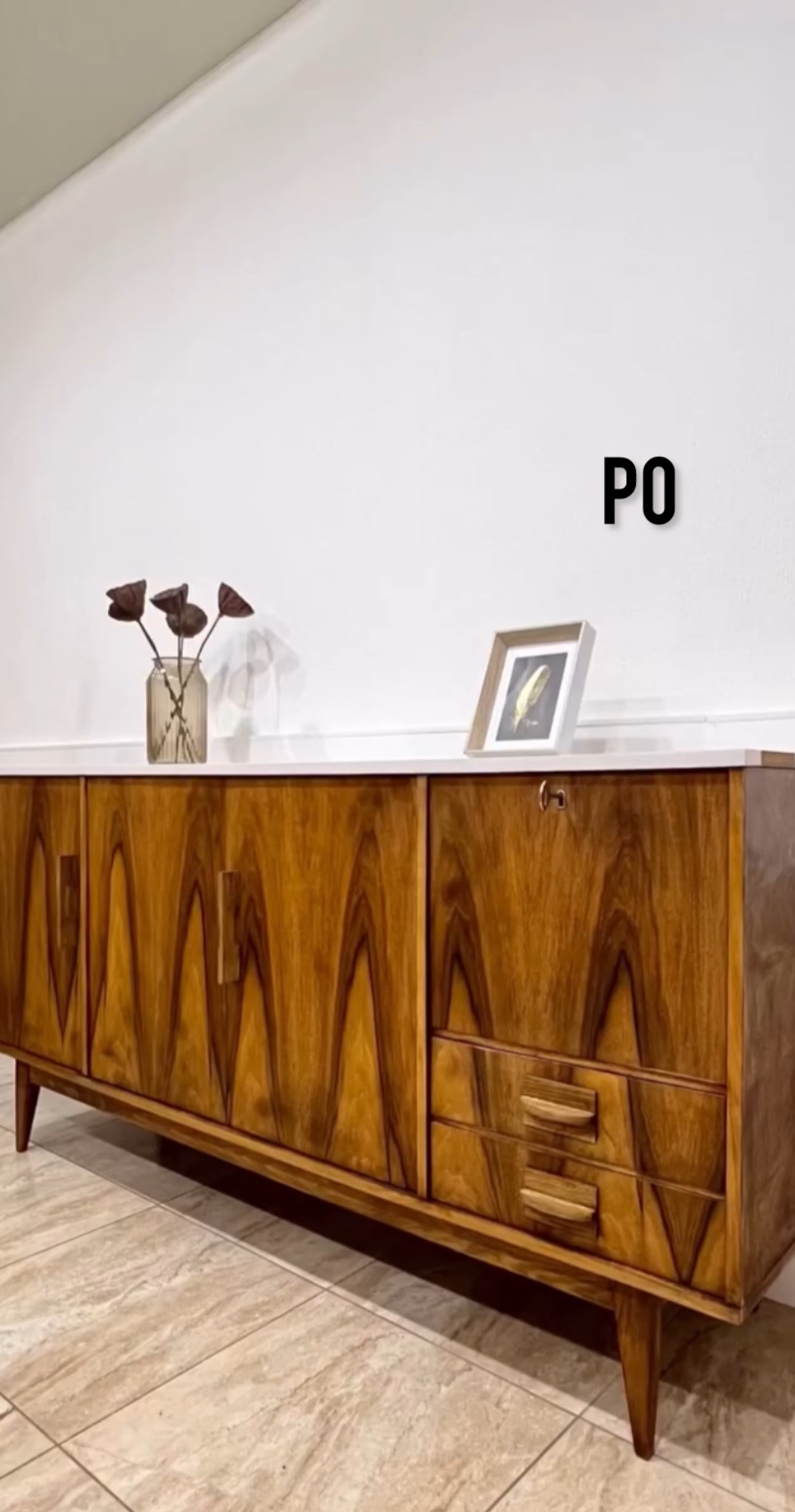 Odrestaurowany kredens z fornirem orzechowym, widoczne detale frezowania frontów i nóżek. Na blacie wazon z suszem i ramka ze zdjęciem. Napis 'PO' sugeruje metamorfozę.