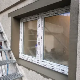 Montaż okna PCV marki Rehau w ościeżnicy z szarą, świeżo nałożoną zaprawą, widoczne metalowe schody z boku.