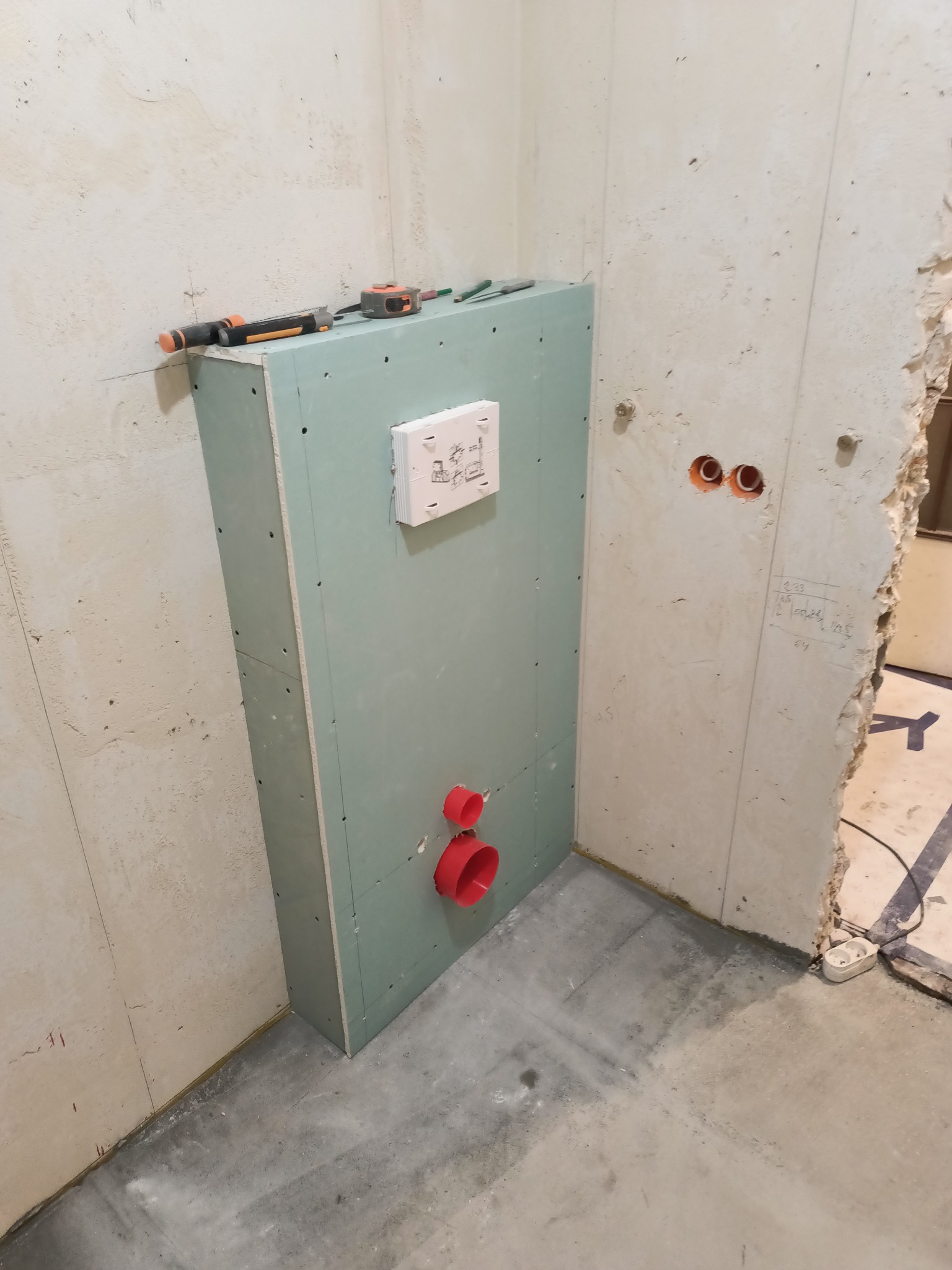 Stelaż podtynkowy obudowany płytą gipsowo-kartonową z widocznymi elementami instalacji wodno-kanalizacyjnej i przyciskiem spłukującym, narzędzia na górnej krawędzi, surowy stan wykończenia wnętrza.