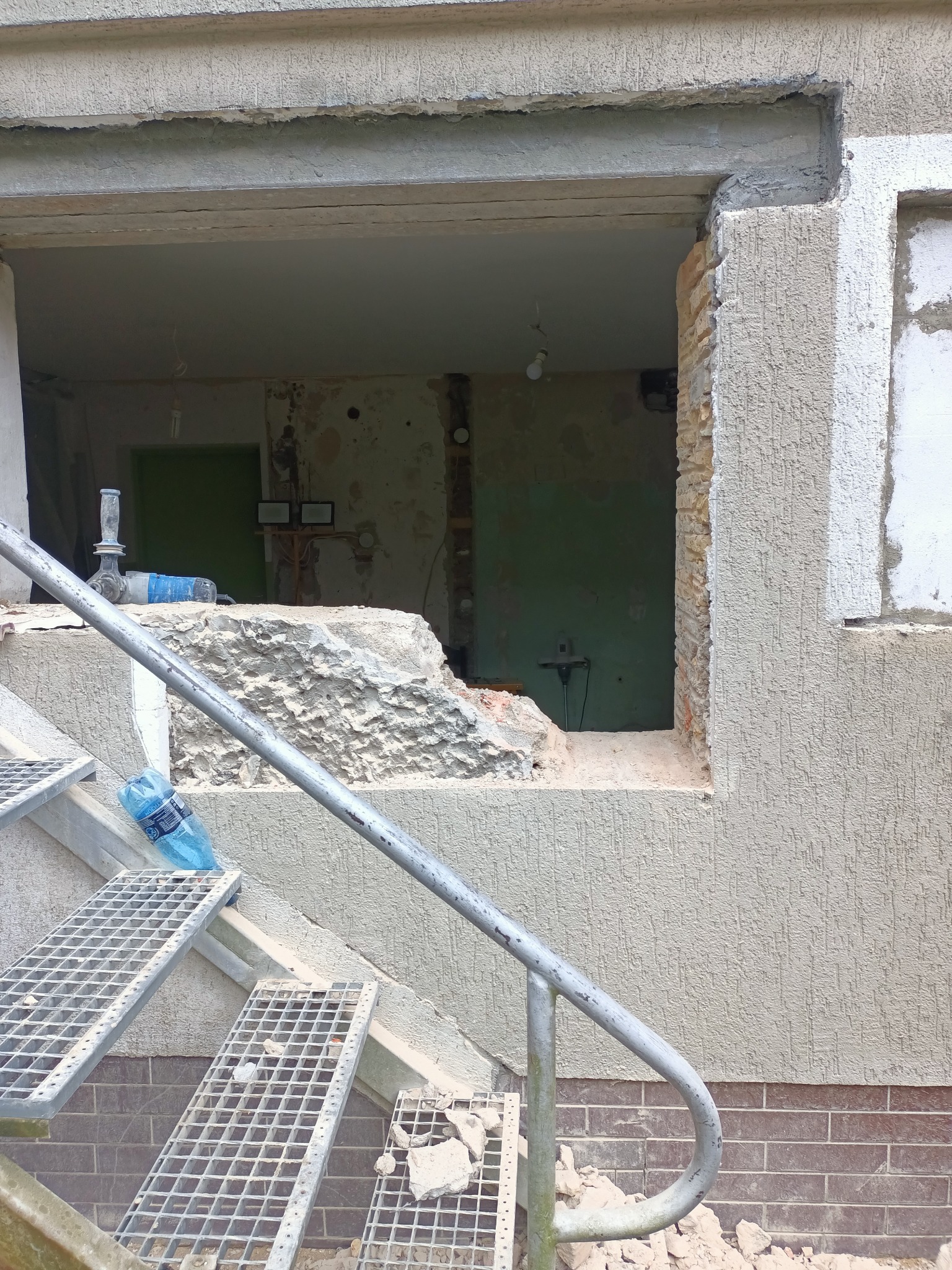 Otwór okienny w trakcie powiększania w elewacji budynku, widoczne narzędzia i butelka wody na schodach.