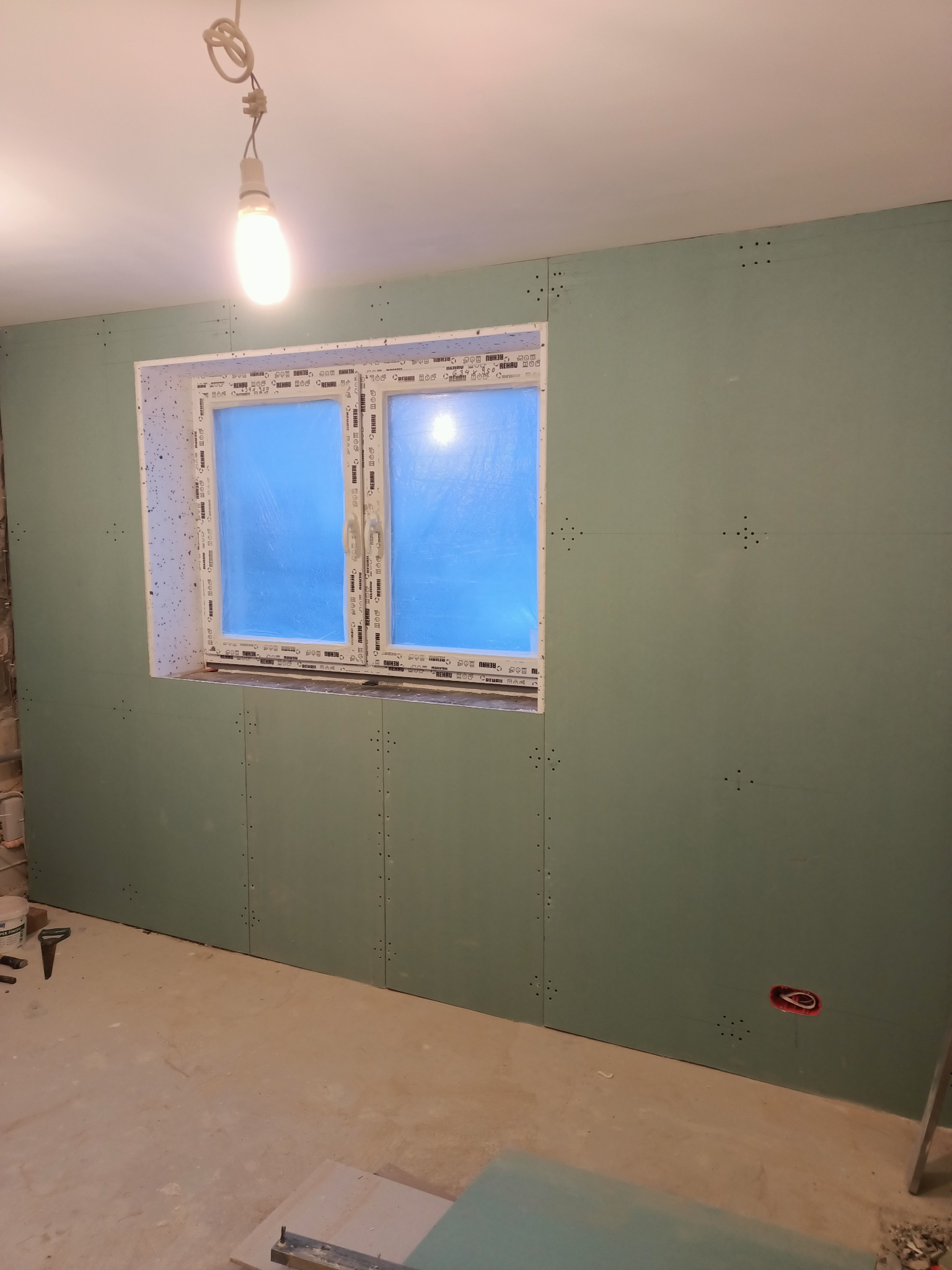 Wnętrze w trakcie wykańczania: ściany pokryte płytami gipsowo-kartonowymi, widoczne łączenia i otwory na wkręty, okno z folią ochronną, surowa żarówka zwisająca z sufitu, widoczne narzędzia...