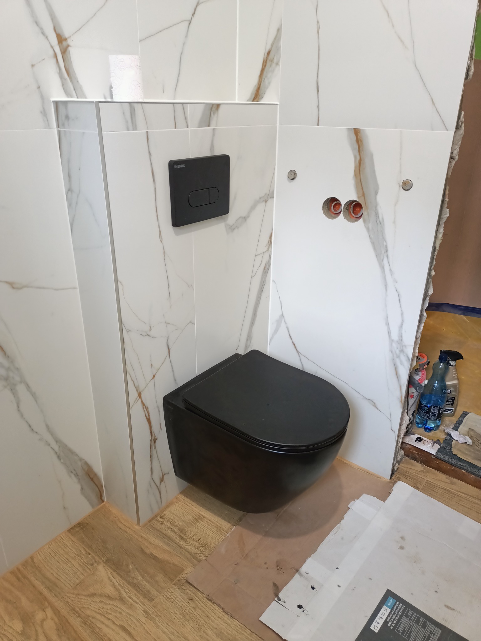 Czarna, podwieszana toaleta z przyciskiem spłukującym na ścianie wyłożonej białymi płytkami imitującymi marmur. Widoczne otwory na instalacje i fragment niezakończonej obróbki ściany z boku.