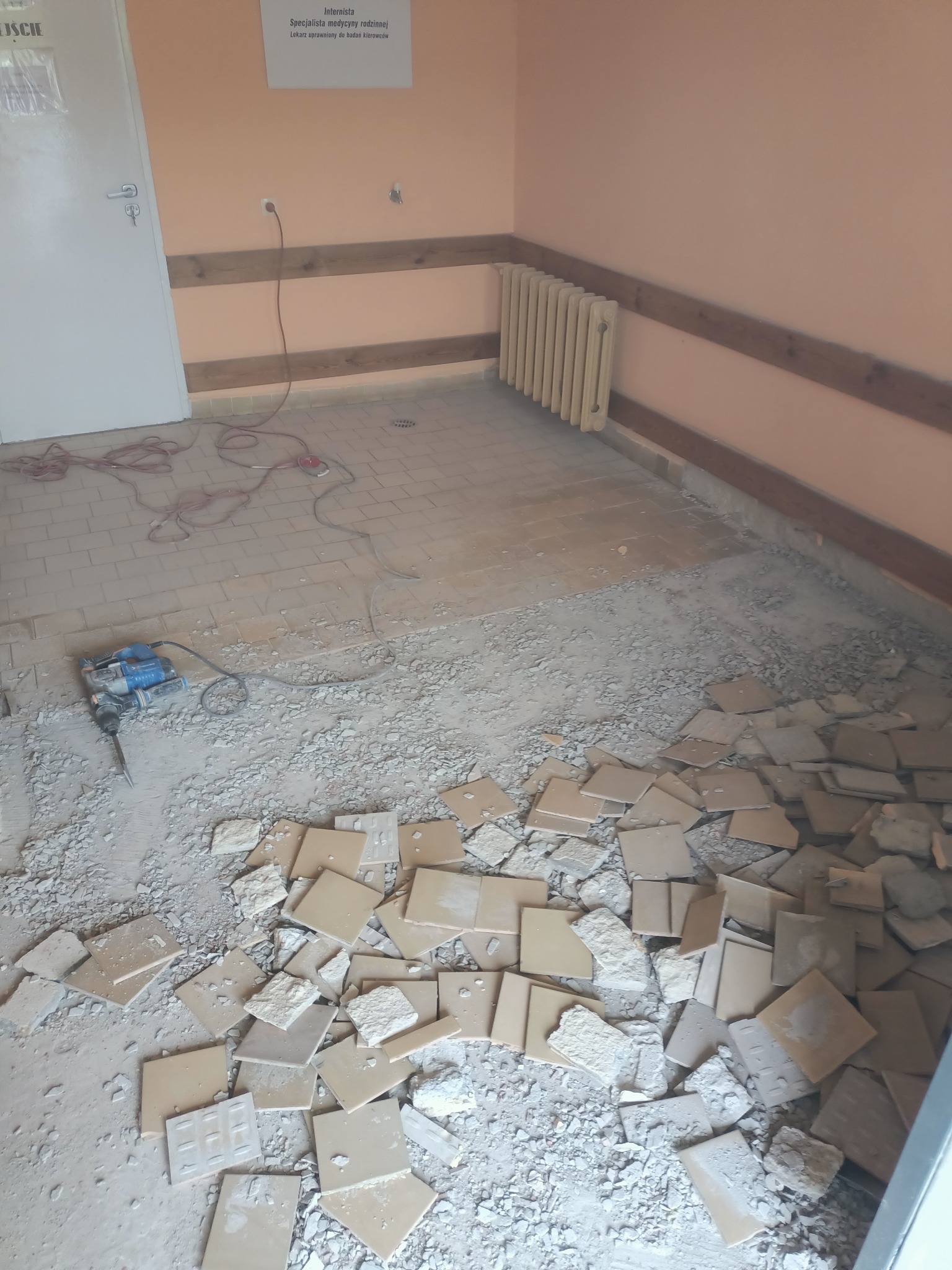 Demontaż starej podłogi z płytek ceramicznych w pomieszczeniu, widoczny gruz, odspojone płytki i młot udarowy