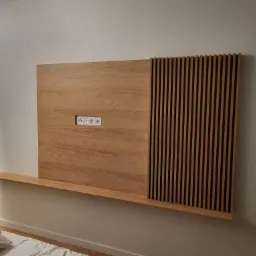 Drewniana płyta ścienna z gniazdkami elektrycznymi i dekoracyjnymi pionowymi listwami, zamontowana na ścianie nad półką.