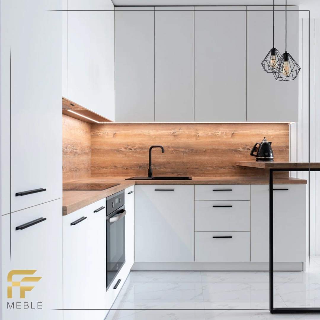 Nowoczesna, jasna kuchnia z białymi frontami i drewnianym blatem oraz panelem ściennym, oświetlona listwą LED, z czarnymi, minimalistycznymi uchwytami i armaturą, uzupełniona czajnikiem...