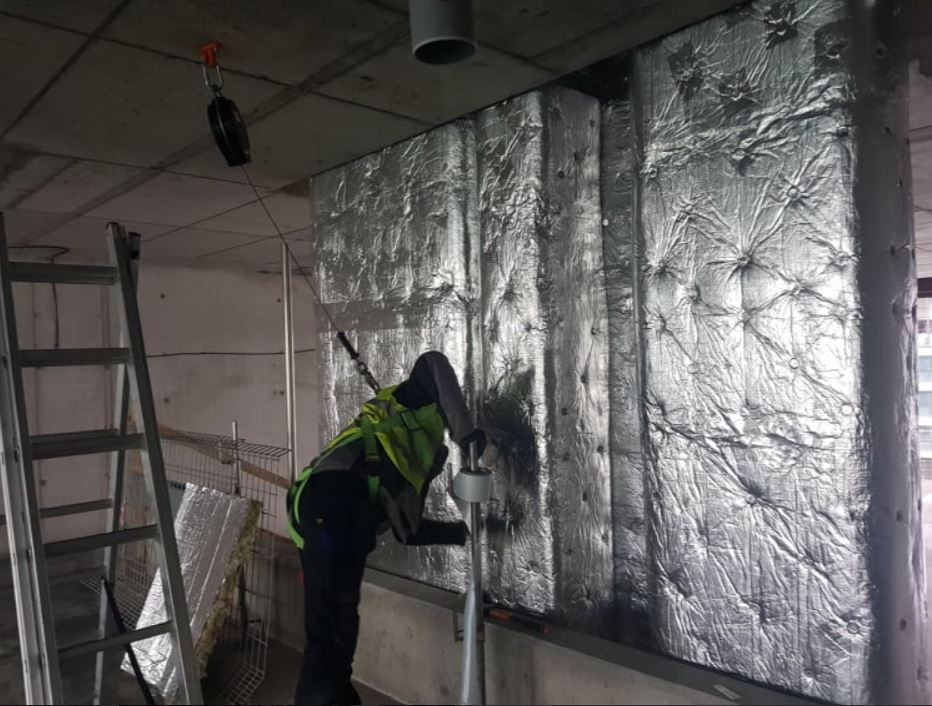Pracownik w kamizelce odblaskowej montuje izolację z folii aluminiowej na ścianie w surowym wnętrzu, widoczna drabina i narzędzia.