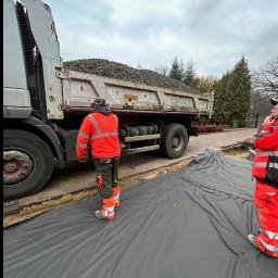 SZAWŁOGA Przedsiębiorstwo Drogowo-Budowlane Eugeniusz Szawłoga - Ciężarówka z kruszywem na budowie, robotnicy w odblaskowych ubraniach przygotowują podłoże wyłożone czarną geowłókniną. Etap prac ziemnych.