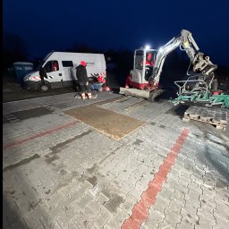 SZAWŁOGA Przedsiębiorstwo Drogowo-Budowlane Eugeniusz Szawłoga - Prace brukarskie w toku: mini koparka z osprzętem, pracownicy, dostawczy samochód. Kostka brukowa częściowo ułożona, widoczne narzędzia i materiały. Praca nocą.