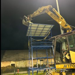 SZAWŁOGA Przedsiębiorstwo Drogowo-Budowlane Eugeniusz Szawłoga - Żółta koparka podnosi platformę na konstrukcji z metalowych rur na tle ciemnego nieba i budynku. Operator w kabinie maszyny. Prace ziemne w nocy.