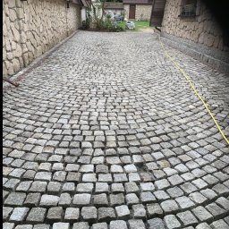 SZAWŁOGA Przedsiębiorstwo Drogowo-Budowlane Eugeniusz Szawłoga - Ułożona z szarej kostki granitowej, zakrzywiona ścieżka między kamiennymi budynkami. Żółty wąż ogrodowy leży wzdłuż ściany po prawej stronie.