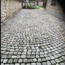 Ułożona z szarej kostki granitowej, zakrzywiona ścieżka między kamiennymi budynkami. Żółty wąż ogrodowy leży wzdłuż ściany po prawej stronie.