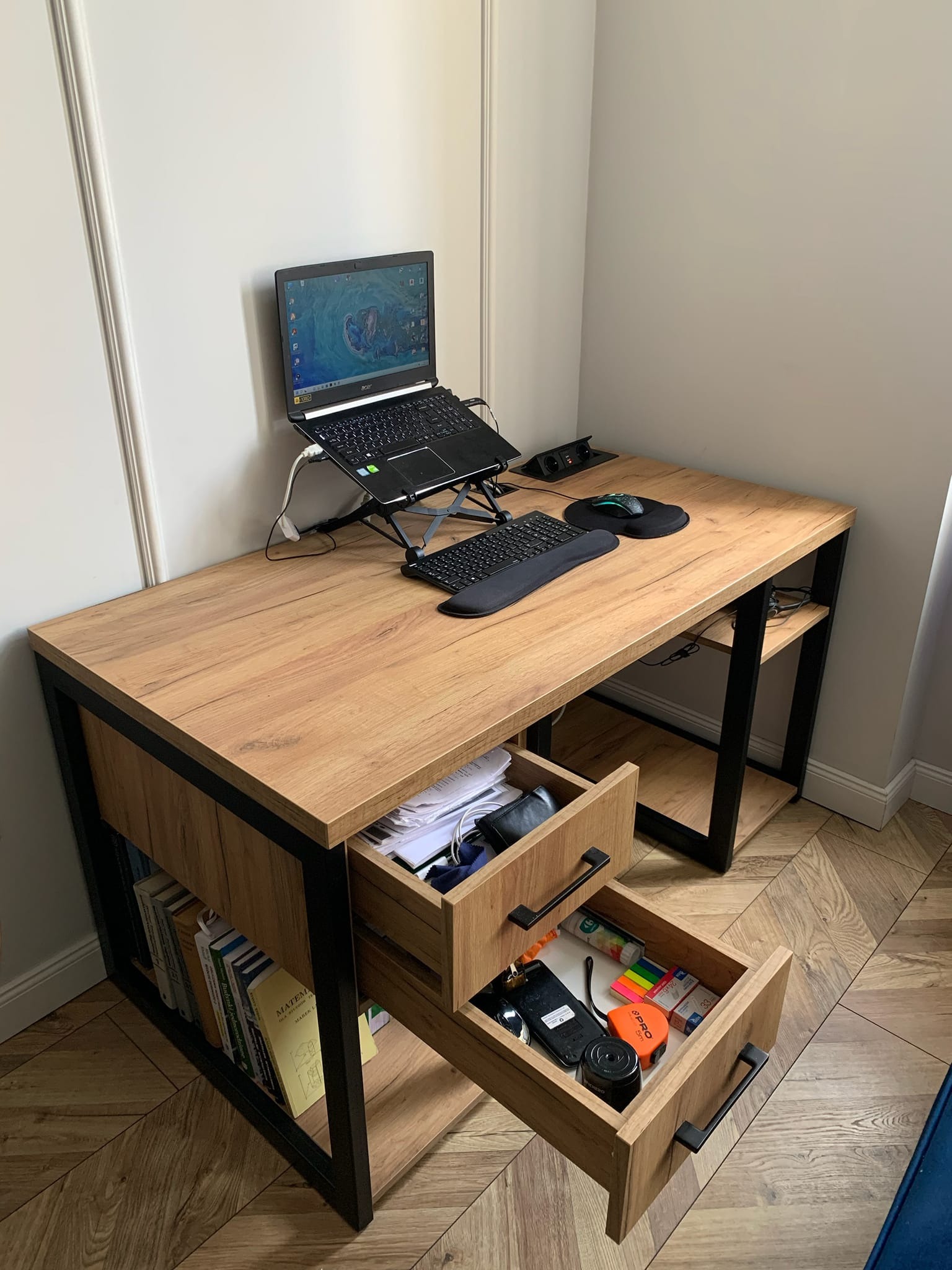 Biurko z blatem imitującym drewno, czarne metalowe nogi, otwarte szuflady z widocznymi akcesoriami biurowymi, laptop na stojaku, klawiatura ergonomiczna, myszka, na półkach książki.