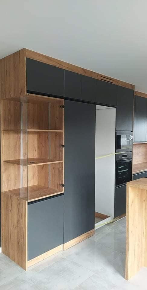 Nowoczesna zabudowa kuchenna z frontami w kolorze grafitowym i elementami z forniru imitującego drewno, obejmująca szafki wiszące, słupek z piekarnikiem i szafkę z przeszklonymi drzwiczkami,...