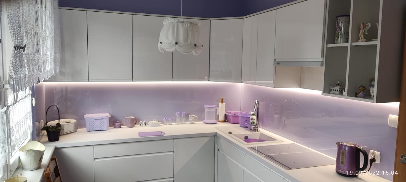 Jasna kuchnia z białymi, błyszczącymi szafkami, fioletowym szkłem lacobel, ozdobną lampą i dopasowanymi kolorystycznie akcesoriami kuchennymi.