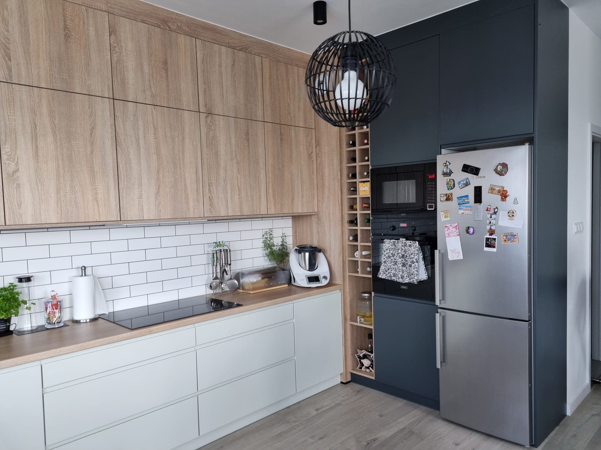Nowoczesna kuchnia z drewnianymi szafkami górnymi, jasnymi szafkami dolnymi, białymi kaflami na ścianie, czarną płytą grzewczą, piekarnikiem i lodówką obok regału na wino oraz ozdobną lampą wiszącą.
