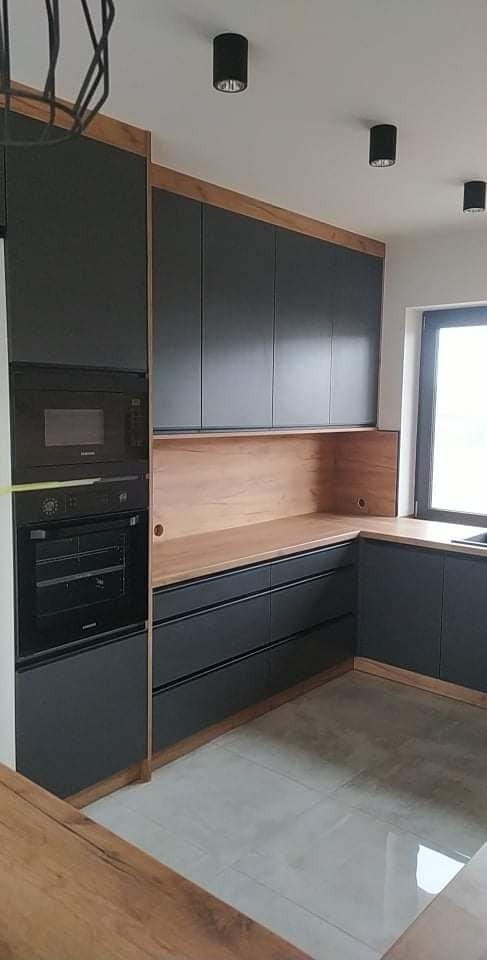 Nowoczesna kuchnia z szarymi szafkami, drewnianymi blatami i wbudowanym piekarnikiem oraz mikrofalówką podświetlona trzema czarnymi lampami sufitowymi.