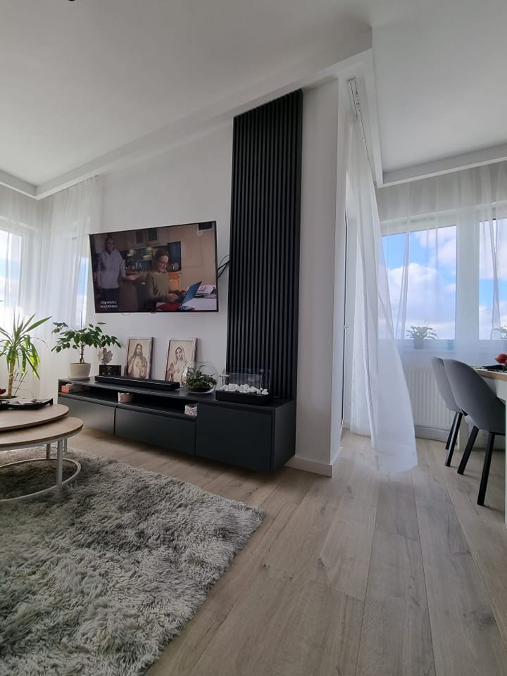 Nowoczesny salon z czarną szafką RTV, pionowym panelem dekoracyjnym i puszystym dywanem w odcieniach szarości, oświetlony naturalnym światłem wpadającym przez okna z białymi firanami.