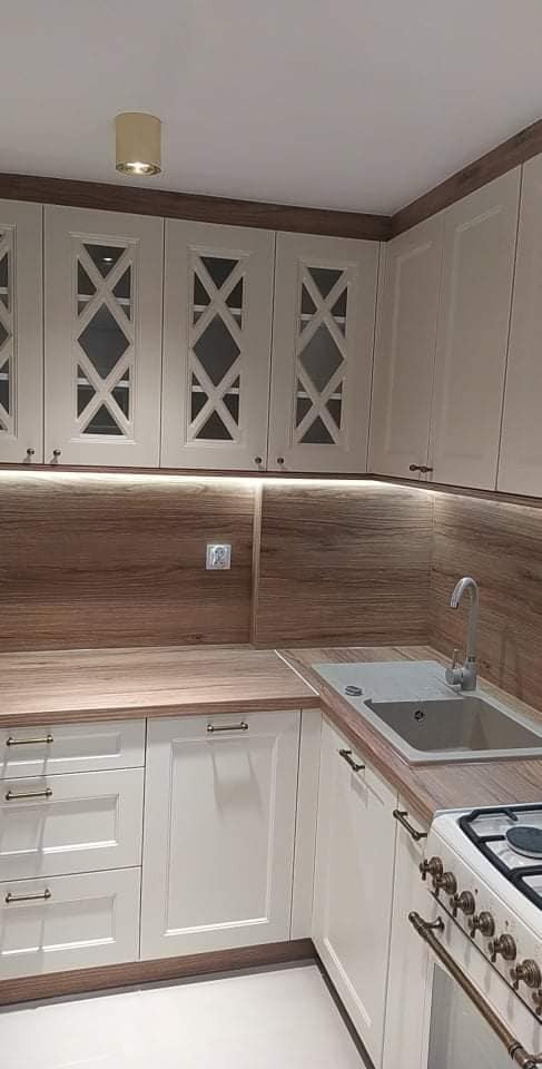 Jasna kuchnia z frezowanymi frontami szafek górnych z motywem rombu, drewnianym blatem i pasującym panelem ściennym, oświetlona listwą LED pod szafkami wiszącymi.