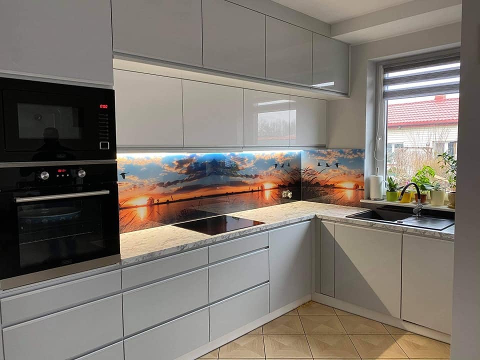 Nowoczesna kuchnia z białymi, lakierowanymi frontami, marmurowym blatem i panelem szklanym z motywem zachodu słońca nad jeziorem, z widocznymi ptakami w locie.