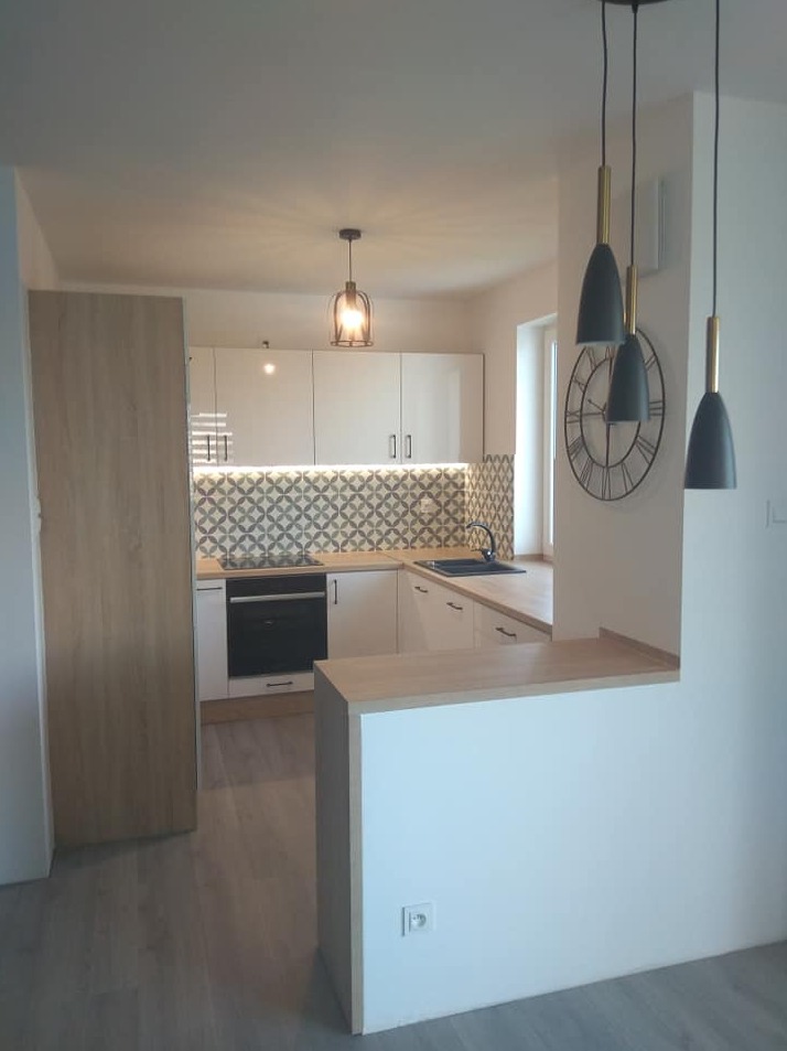 Jasna kuchnia z białymi szafkami, drewnianym blatem i wzorzystą mozaiką nad blatem, oświetlona trzema wiszącymi lampami w stylu loft oraz pojedynczą lampą sufitową, widoczny zegar na ścianie.