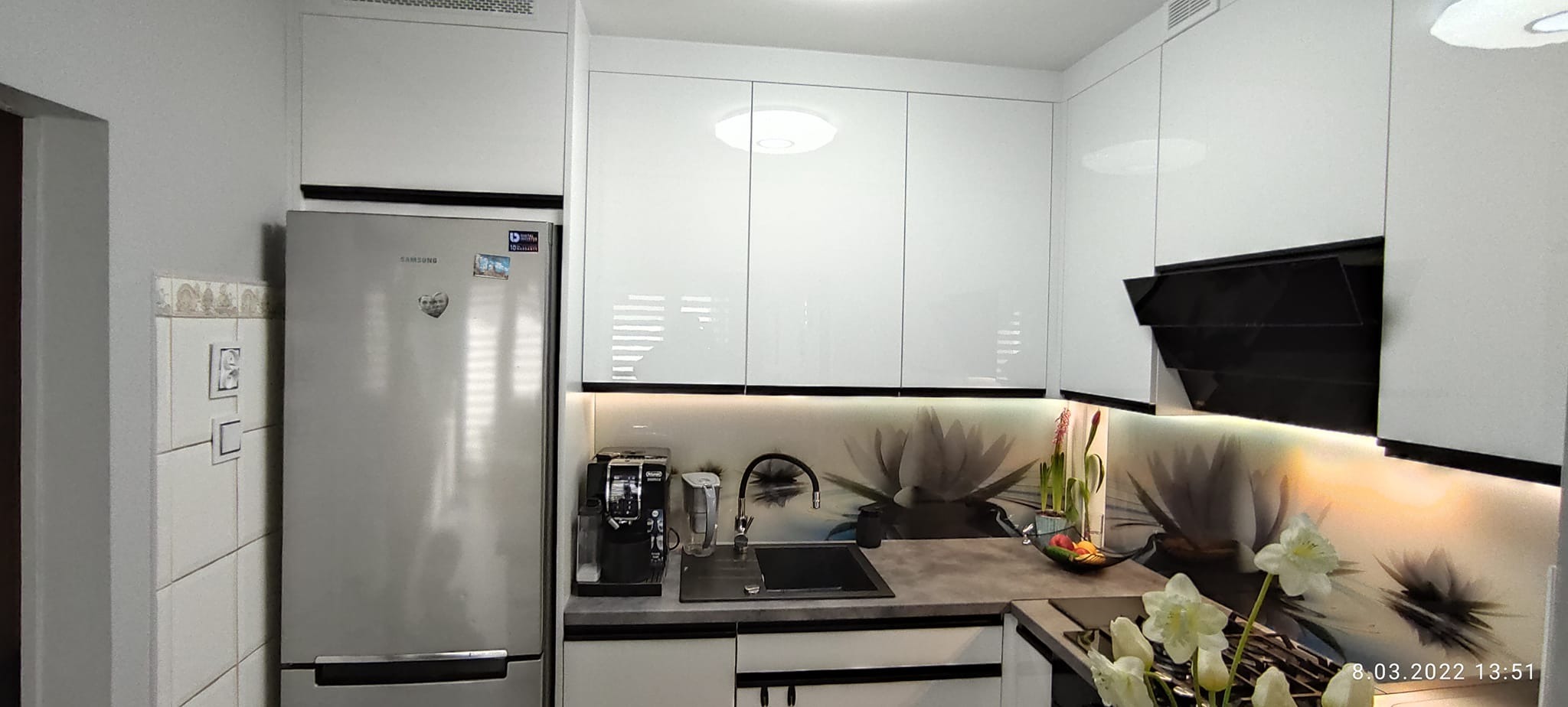 Nowoczesna kuchnia z białymi, lakierowanymi szafkami, czarnym okapem i blatem, z fototapetą z motywem kwiatowym, oświetleniem LED pod szafkami, srebrną lodówką Samsung i ekspresem do kawy.