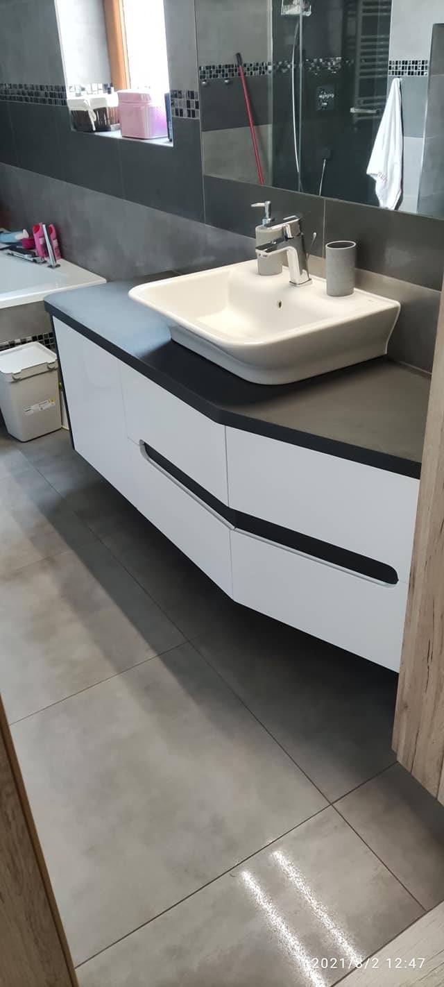 Nowoczesna łazienka z białą umywalką nablatową na szafce z czarnymi akcentami, szare płytki podłogowe i fragment lustra z widoczną baterią.