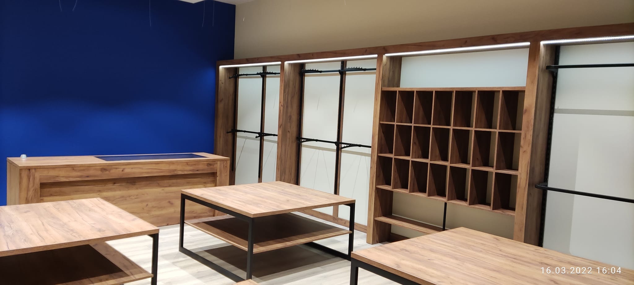 Wyposażenie sklepu odzieżowego: drewniane regały z oświetleniem LED, wieszaki na ubrania, stoliki z metalowymi nogami i drewnianym blatem, lada z ciemnym blatem, ściana pomalowana na intensywny...