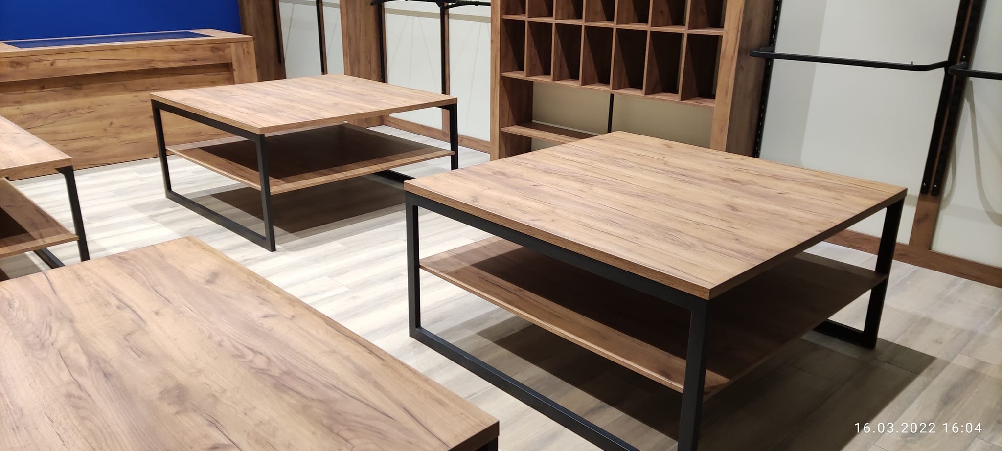 Komplet drewnianych stolików kawowych z czarnymi, metalowymi nogami i półkami w sklepie odzieżowym, na podłodze jasne panele, w tle regał z przegródkami i wieszaki na ubrania.