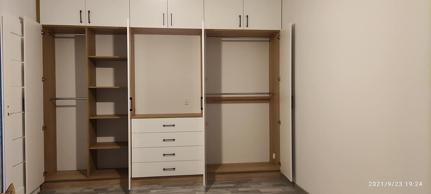 Biała szafa wnękowa z drewnianymi elementami, częściowo otwarta, ukazująca półki i szuflady, z widocznym fragmentem podłogi