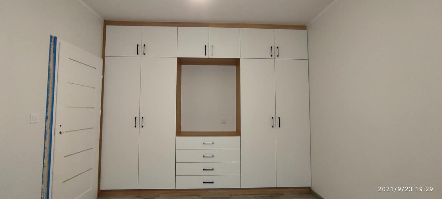 Biała szafa wnękowa z drewnianymi akcentami i czarnymi uchwytami, wbudowana w ścianę, widoczna również biała ościeżnica drzwi.