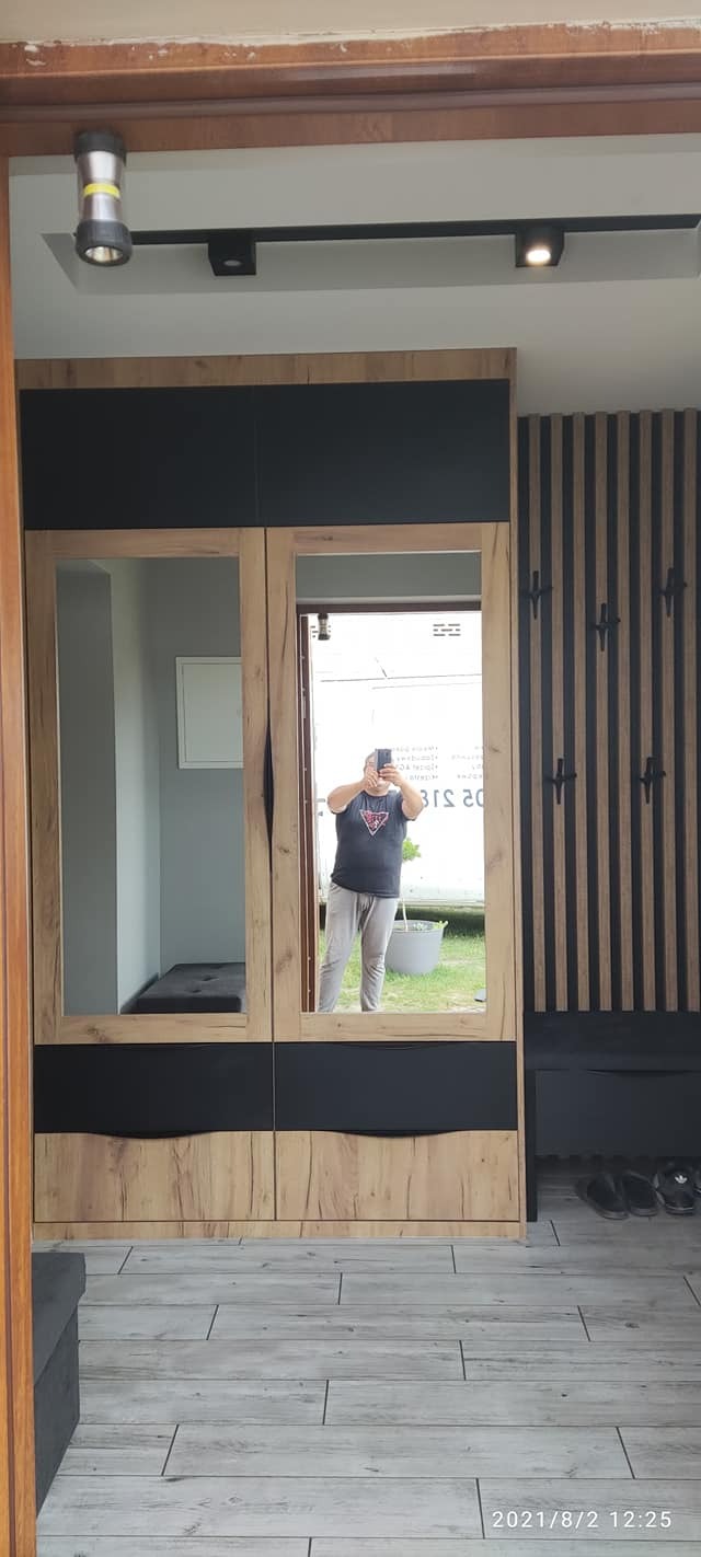 Wnętrze przedpokoju z szafą wnękową z lustrami, drewnianymi panelami ściennymi i siedziskiem, oświetlenie sufitowe punktowe, odbicie osoby robiącej zdjęcie w lustrze.