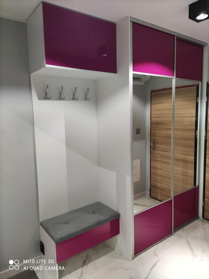 Biała szafa wnękowa z przesuwnymi drzwiami lustrzanymi i fuksjowymi akcentami, wbudowane siedzisko z szarą poduszką i wieszakami na ubrania.