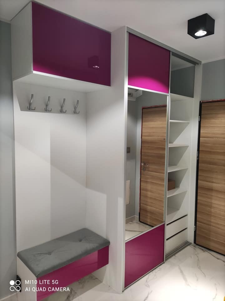 Wnęka z szafą przesuwną, siedziskiem i wieszakami, wykończona w bieli i fuksji, z lustrzanym frontem i drewnianymi drzwiami w tle.